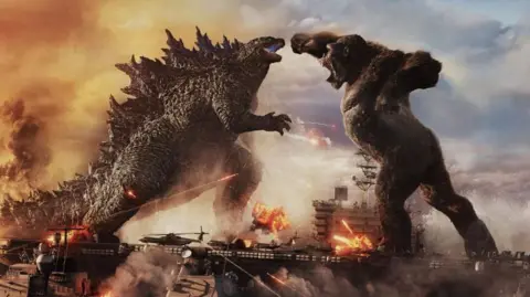 WarnerBros.com A still from the Godzilla vs Kong movie. The two titular monsters engage in battle while standing on military battle ships.