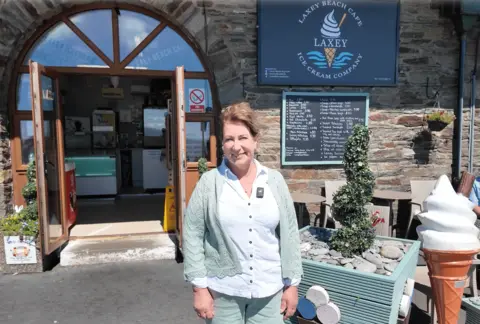 Julie Pinson, wearing a teal cardigan and white shirt, stands in front of Laxey Beach Cafe. In the background you can see a large plastic ice cream, and a swirling garden tree. 