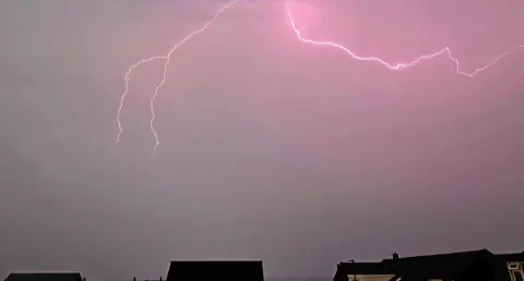 Sharon Bell Rooftops silhouetted by pink lightning strikes above grey thundery skies. 