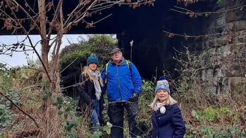 Two blonde women and a man stand in overgrown greenery under a railway bridge