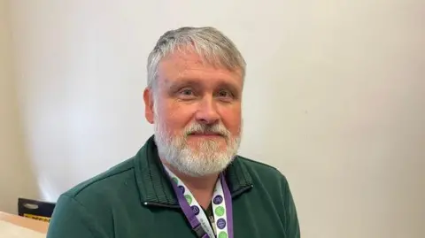 Trevor Lucy Trevor has a well-kept grey beard and grey hair with short sides and slightly longer fringe. His fleece is dark green around neck are lanyard ribbons.