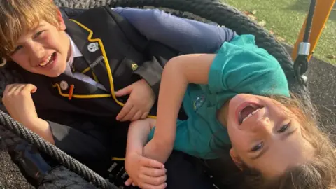Gordon Hunt Henry y Holly ríen mientras están sentados juntos en un gran columpio circular de cuerda en un parque infantil. Henry lleva uniforme escolar y Holly una camiseta verde, vistos desde arriba mientras el columpio se inclina.
