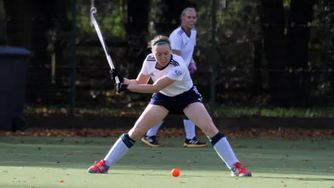 Chichester Hockey Club A women's player in a white kit swinging a hockey stick at an orange ball