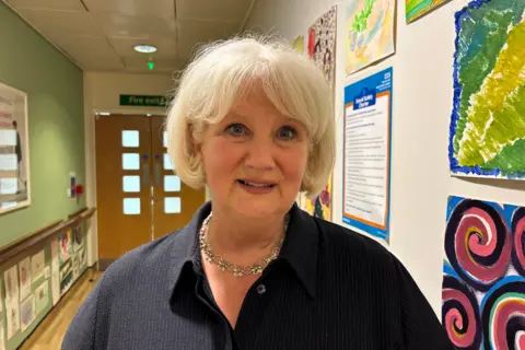 Woman with white bobbed hair, navy top, and a gold intricate necklace, standing in a corridor which has paintings on the righthand side, and then a green wall on the left and doors at the end of the corridor.