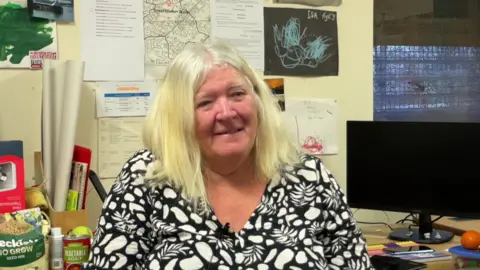Spencer Stokes/BBC A woman in a black and white flowery top, sitting in an office, smiles at the camera.