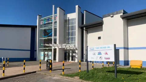 The Dumfries Ice Bowl with a white and blue facade and glass entrance way. There are black and yellow bollards outside, a sign saying what teams play there and a black bin.
