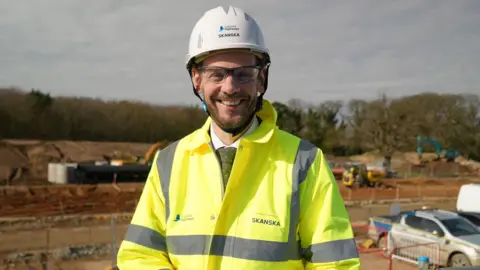 Shaun Whitmore/BBC Simon Lightwood is grinning at the camera. He is wearing glasses and a white hard hat and high-vis jacket both branded with Skanska. He has a white shirt and green spotted tie underneath. He is standing in front of a construction site.