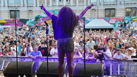 A woman is standing on a stage seen from the back. A crowd is facing the stage.