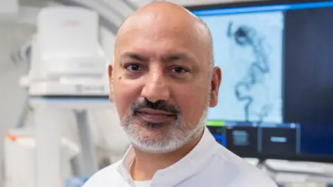 A man with short white beard wearing a white shirt stands in a medical room