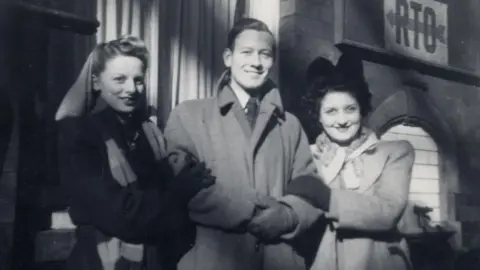 Family handout A black and white image of Carol with friends outside a theatre after a show. Carol is on the left wearing a dark coat and grey and black chunky scarf. She has her hair half up and half down. A man in the middle is wearing a grey overcoat with a shirt and tie beneath. The woman on the right is wearing a big black bow in her hair and a light-coloured overcoat and scarf.