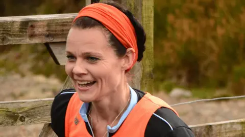 Michael Gregory Emma Gregory running on fells, wearing an orange headband and orange running vest, with a black tshirt. She has her dark hair tied back and is smiling as she is mid-stride.