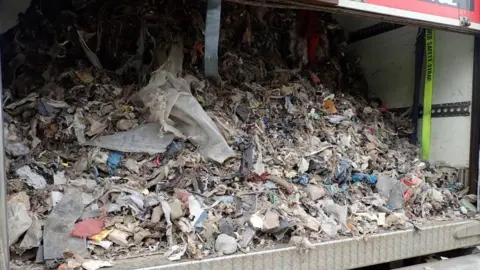 Rear of a trailer with the roller door partly open, showing it is filled with fist-sized pieces of dry waste mixed with plastic and textile. 