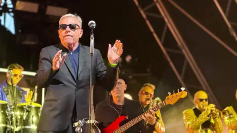 Madness on stage. Suggs is clapping his hands while musicians play behind.