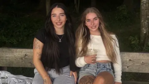 Two girls sitting on a wooden park bench. The one on the left is wearing a black t-shirt and jeans. She has dark brown hair. The girl on the right is wearing a white jumper and blue shorts. She has light brown hair.
