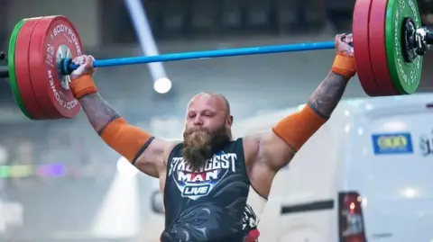 A man in a dark leotard, with a bald head and long brown beard is holding a huge weight above his head. His leotard has the words "UK Strongest Man Live" printed on it.