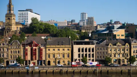 Multi-storey old-style light-coloured buildings with several windows along a riverfront with a church in the background. 