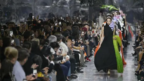 Getty Images Models walking down a mirrored runway, the first wearing a black draped sleeveless dress and holding a lime green fringed bag - people watching the show are applauding, many of them holding up their phones to take pictures 