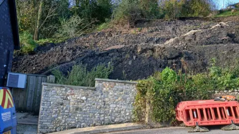 Dorset Council A collapsed bank next to a steep road. The collapse is behind a stone wall but the bare earth rises up behind it and appears to be about three times as high at the wall.