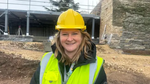 Sarah Clarke in a high vis jacket and hard hat, stood outside the castle walls, looking at the camera