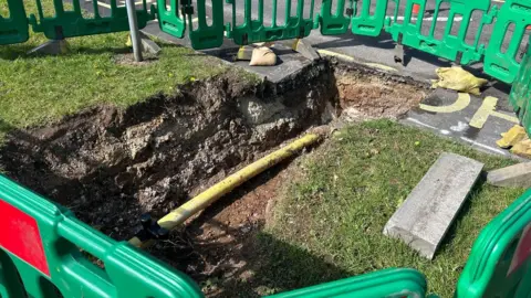 BBC A yellow gas pipe in a deep trench, which has been dug so as to remove part of the word "school" that was previously painted on a road
