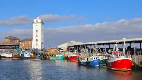 Figaro About seven fishing boats moored in front of a of warehouse with a corregated roof. Vehicles are parked under the roof. To the left are brown brick houses and a tall, white tower with a look-out deck. The water is calm and the sky is largely blue with some white, grey clouds.