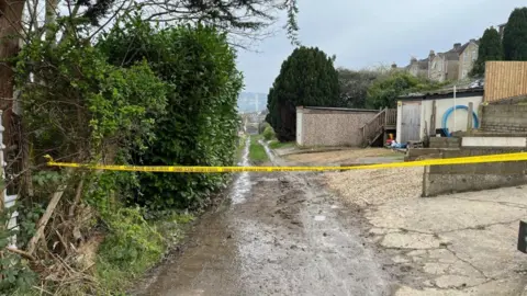 BBC Police tape stretching across a muddy footbath in Bath, with trees and outbuildings visible