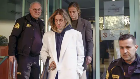 EPA Alexia Putellas and Irene Paredes leave court. Two security guards are next to them, one of them holding open a glass door for the two women. One of the women is wearing a white coat and has blonde hair, and the other is wearing a dark brown blazer and has brown hair.