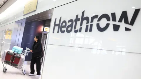 A woman walks through a doorway pushing a trolley with luggage stacked on it with the word Heathrow written in black beside her