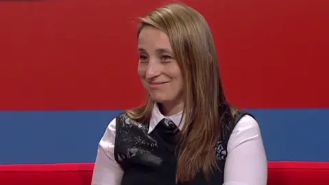 A woman with long brown hair sits on a sofa in a TV studio and smiles. She wears a white shirt with a black sleeveless shirt on top of it.