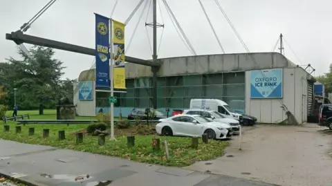 Google Street View of Oxford Ice Rink's exterior, a large white and blue building, with some moss growing on the roof. Large windows are at its centre.