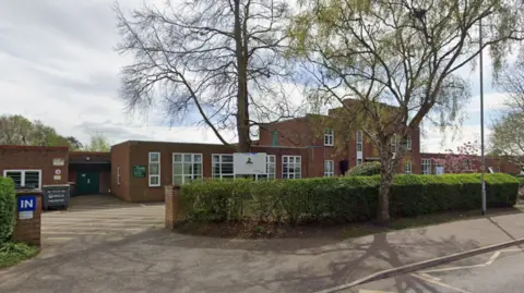 Google The outside of Firside Junior School in Norwich. A red-brick building with a hedge in front of the site enclosing a car park. 