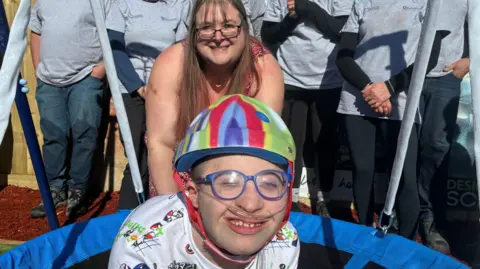 Image of a teenage boy wearing a multi-coloured helmet, blue glasses and with an oxygen line smiling at the camera. In the background can be seen a woman in a summer dress and glasses, who is leaning forward and smiling.