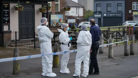 JMG Press Three forensics officers talking to a man dressed in black trousers and navy shirt at a police cordon around a pub.