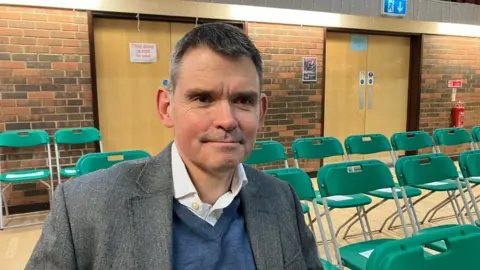 Amy Holmes/BBC A man sits on a green folding chair in a hall, wearing a grey blazer over a blue sweater and white shirt, with rows of similar chairs and closed wooden doors in the background.
