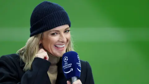 Richard Sellers/Allstar A blonde-haired woman with a happy face, wearing a beige turtleneck, black jacket and black hat smiling and looking off  into the distance while holding a microphone.  