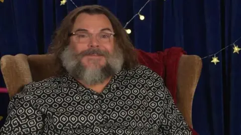 A man with brown hair, glasses and a grey beard is sitting in a brown chair. He's wearing a black and white shirt, and behind him is a blue curtain and fairy lights