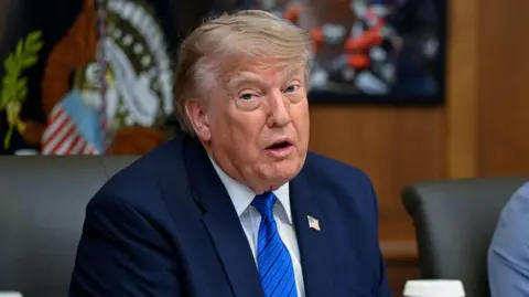 Donald Trump sitting at a US cabinet meeting at Camp David on 31 July, wearing a dark blue suit, a lighter blue tie with black horizontal stripes, a white shirt and a badge of the US flag pinned to his jacket.
