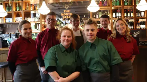 TEAM DOMENICA The image shows a group of seven people posing: the two people at the front are wearing dark green shirts while the rest are wearing red. They are stood in a pub with a sign reading "The North Star" behind them on a mirror. Bottles of alcohol can be seen on shelves in the background.