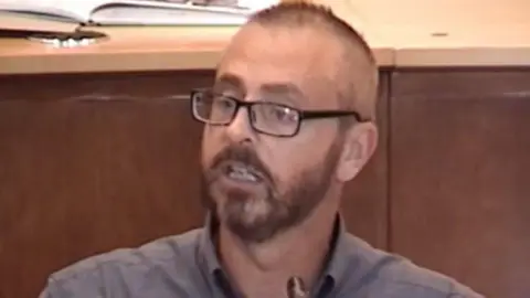 LDRS A blurry image of a man with short dark hair and a beard, wearing glasses and a grey shirt.