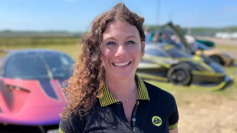 Sheena Valentina a woman with brunette curly hair is standing outside in front of two motorsport cars. She is looking directly at the camera and is smiling. She is wearing a black T-shirt with yellow detailing which also has a yellow Lotus logo on it.