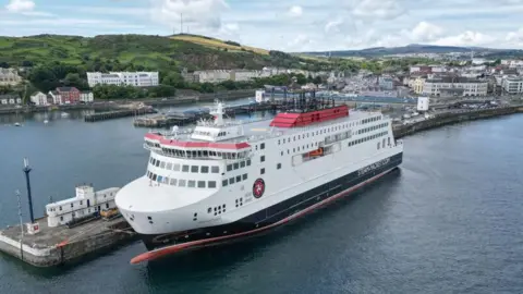 An arial view of Manxman, a white, navy and red ferry, which is moored on the Douglas outer harbour, you can see hills and buildings in the background.