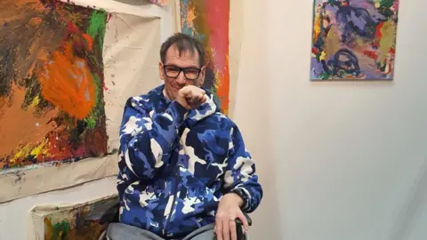 Grundy Art Gallery Stephen Crawford has a multi-cloured hoodies and is sitting in front of some of his work.