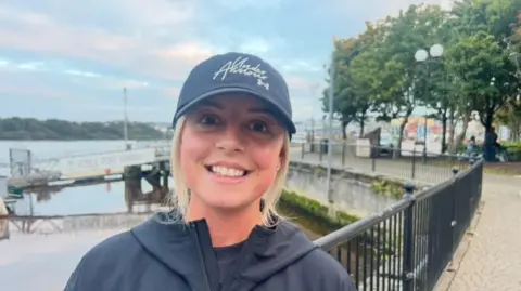 Natasha Weir in dark grey jacket and cap, with blonde hair, beside railings and the river.