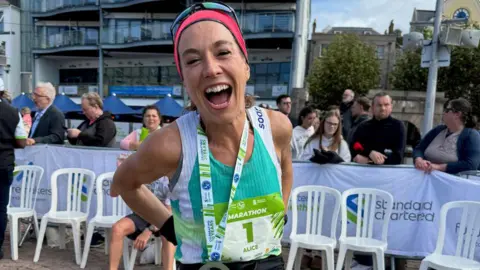 Alice Braham Seorang wanita tersenyum lebar, mengenakan bib nomor 1 untuk Jersey Marathon, dengan medali di lehernya dan orang-orang di belakang penghalang di belakangnya.
