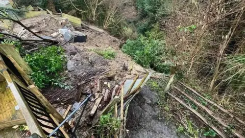 A view of the landslip from a back garden affected which shows the downward slope covered in mud as well as fallen fences, debris and shrubs with more trees at the bottom of the slope.