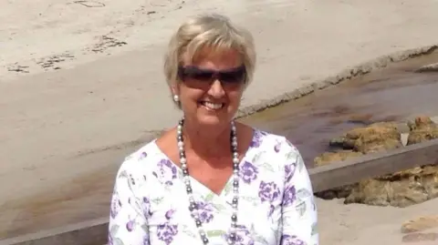 Ruth Smith Mrs Smith wearing a white and purple floral blouse and sunglasses. She has short blonde hair