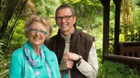 Four Seasons Garden Married couple Marie and Tony stand next to each other in a lush green garden, with the back drop of lots of foliage. Marie is wearing a jade green top and matching patterned scarf. She is wearing green-rimmed glasses and smiling at the camera. Tony is wearing brown jeans and gilet, with a stone coloured long sleeved top, holding a garden tool/broom. He is wearing brown-rimmed glasses and is smiling at the camera. 
