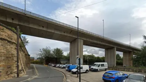 Google The small car park on Pottery Lane. A number of vehicles are parked in the car park, which sits underneath a large concrete bridge. Pottery Lane is on the left-hand side of the picture and runs alongside a high brick wall.