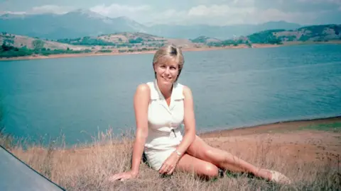 BBC Jill Dando sits smiling at the camera beside a lake in Italy in a promotional photo for the TV series Holiday. She wears a white sleeveless outfit. Large hills can be seen on the other side of the water.