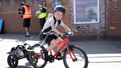 A smiling Henry cycling on his red bike with his ventilator in a trailer behind. He is wearing a black helmet, a white shirt with blue stripes and black shorts.

